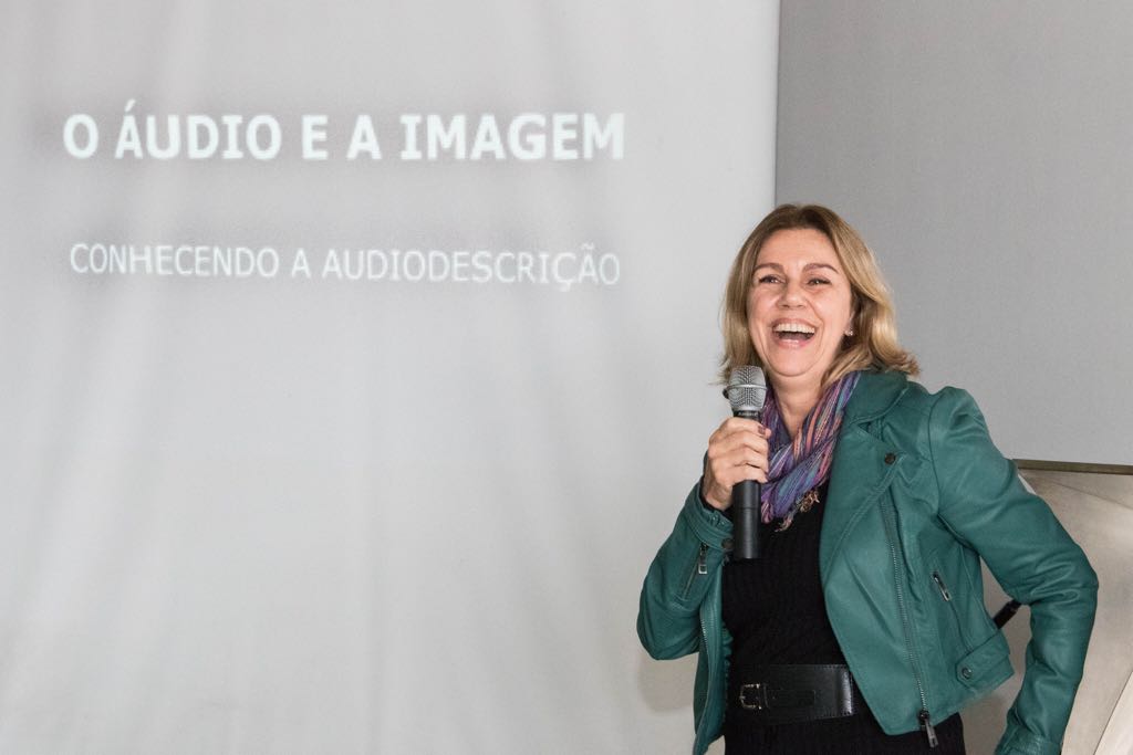 Marisa Pretti sorridente segura o microfone em palestra: o áudio e a imagem, conhecendo a audiodescrição. 