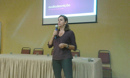 Palestra Faculdade São Camilo: Marisa em pé, a frente da bancada com cadeiras, segura o microfone, ao fundo telão com Power Point da apresentação. 